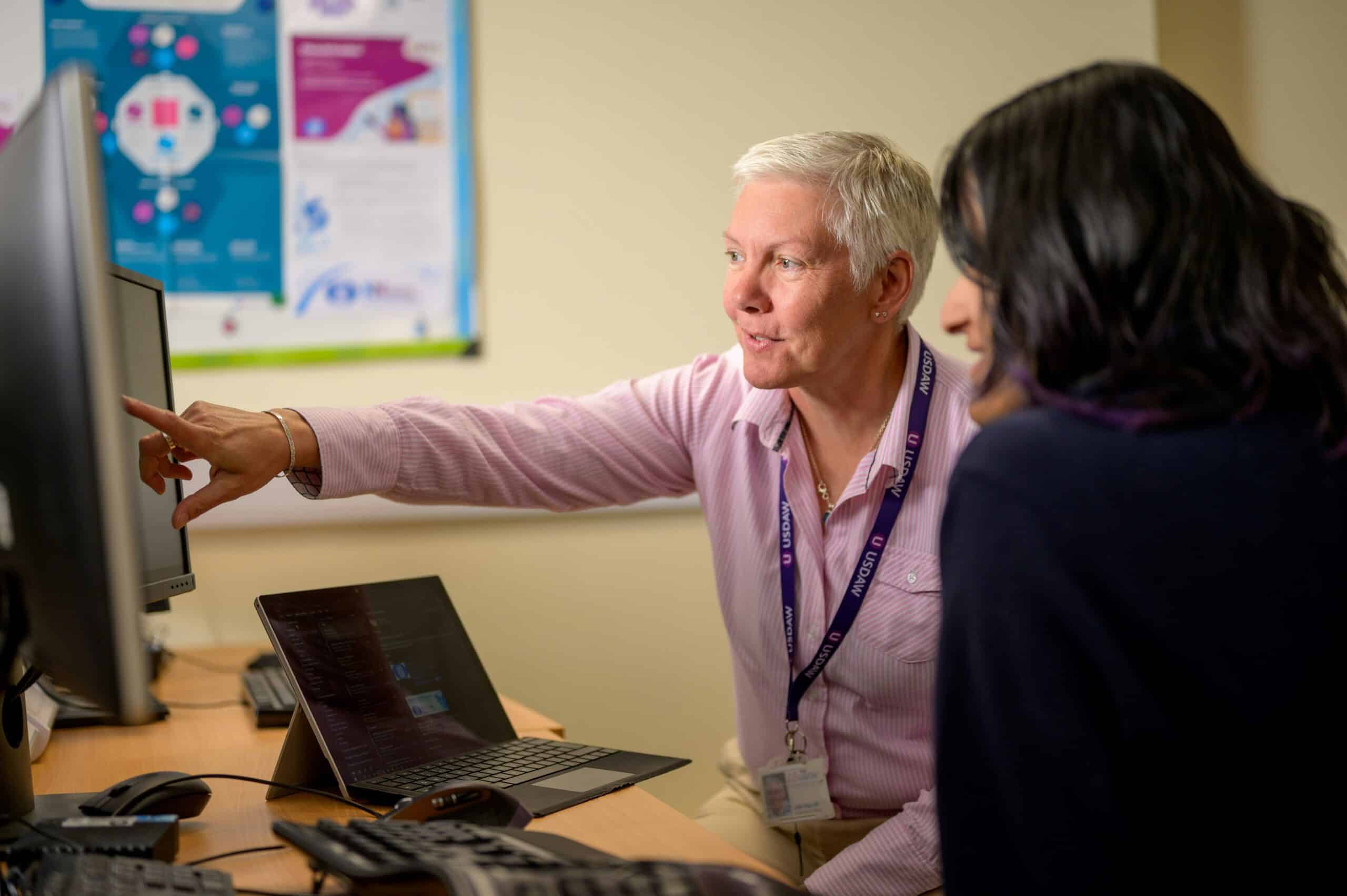 Two women at work talking together at a desk. One is pointing at the computer screen.